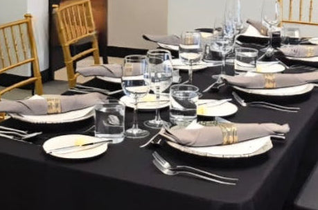 Elegant private dining setup with black tablecloth, neatly arranged plates, glasses, silverware, and folded napkins on a refined dining table with wooden chairs.